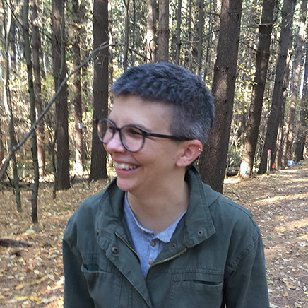 Nikki Finch wearing a green button up shirt, standing in front of a tree and laugh.