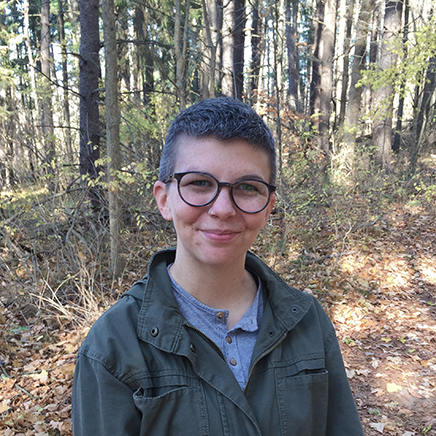 Nikki Finch wearing a green button up shirt, standing in front of a tree and smiling.